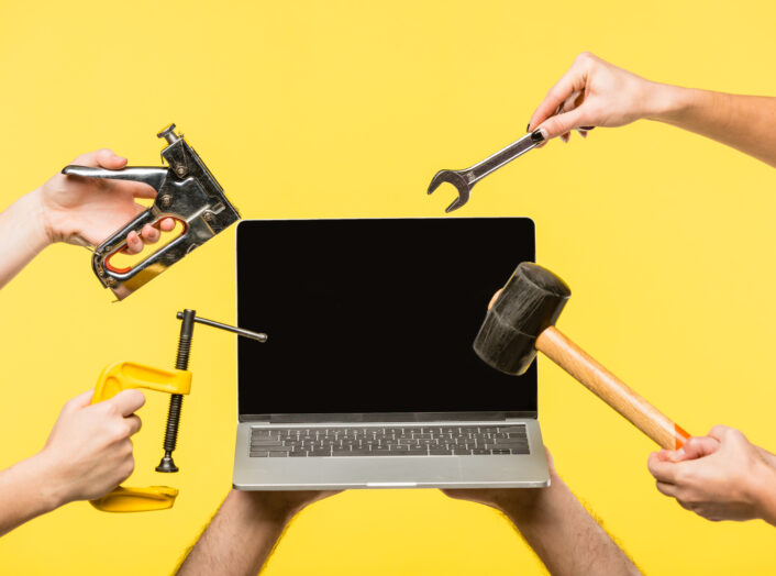 cropped shot of hands holding tools and laptop with blank screen isolated on yellow